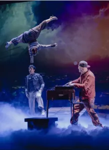 Ein Akrobat vollführt eine spektakuläre Balanceübung auf dem Kopf eines Partners, während der Musiker Supa-Jay live auf der Bühne spielt. Dies veranschaulicht die Mischung aus zeitgenössischem Zirkus, Live-Musik und Video in der Show Sans regrets?.