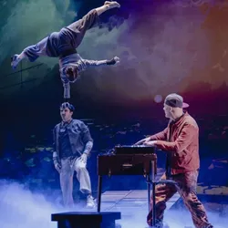 Ein Akrobat vollführt eine spektakuläre Balanceübung auf dem Kopf eines Partners, während der Musiker Supa-Jay live auf der Bühne spielt. Dies veranschaulicht die Mischung aus zeitgenössischem Zirkus, Live-Musik und Video in der Show Sans regrets?.