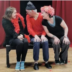 Drie deelnemers aan een clownworkshop, zittend op een bankje voor een rood gordijn, wisselen een veelbetekenende blik uit met hun rode neuzen en kleurrijke kostuums tijdens een workshop georganiseerd door de vereniging Culture et Loisirs Jean Macé.