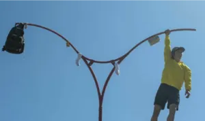 Onder een blauwe hemel hangt Mélusine Lavinet-Drouet, gekleed in een geel jasje, zwarte korte broek en een rode pet, met één hand aan het uiteinde van een grote rode metalen constructie. Haar lichaam zwaait de leegte in, haar benen gebogen en haar arm vri