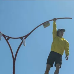 Onder een blauwe hemel hangt Mélusine Lavinet-Drouet, gekleed in een geel jasje, zwarte korte broek en een rode pet, met één hand aan het uiteinde van een grote rode metalen constructie. Haar lichaam zwaait de leegte in, haar benen gebogen en haar arm vri