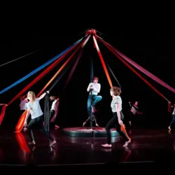 Deelnemers van alle leeftijden bewegen zich tijdens een groepsworkshop rond een circuspaal die versierd is met lange, gekleurde strepen. Dit illustreert de vriendelijke sfeer van het familiecircus tijdens de circusavond.