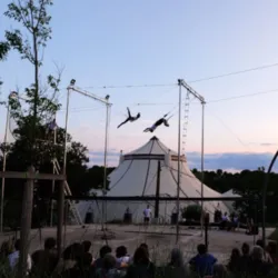 Foto van een grote trapezesessie georganiseerd bij CIAM in Aix-en-Provence. Een trapezeartiest hangt aan de trapeze boven een groot veiligheidsnet, onder een enorme overdekte constructie die natuurlijk licht doorlaat. Deelnemers