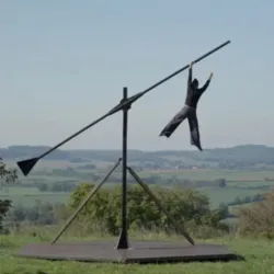 Een acrobaat hangt aan een gigantische metalen windturbine die in evenwicht is in een natuurlijk landschap tijdens de luchtacrobatiekshow "Beyond the Horizon" van Compagnie La Migration.