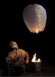 In een nachtelijke atmosfeer, badend in oranje licht, overpeinst Nicolas Fraiseau een lantaarn die opstijgt boven een laaiend vuur. Deze spectaculaire foto uit de IGNIS-tentoonstelling legt de poëzie van vuur vast en combineert magie, hedendaags circus en