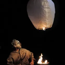 In een nachtelijke atmosfeer, badend in oranje licht, overpeinst Nicolas Fraiseau een lantaarn die opstijgt boven een laaiend vuur. Deze spectaculaire foto uit de IGNIS-tentoonstelling legt de poëzie van vuur vast en combineert magie, hedendaags circus en