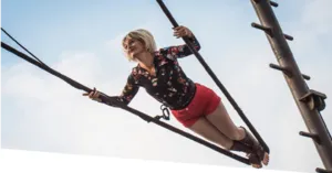 Een foto vanuit een laag perspectief van een blonde artieste in een bloemenblouse en rode korte broek, die hangt en balanceert tussen verschillende kabels die aan een monumentale metalen constructie zijn bevestigd. De foto illustreert La Promesse du Vide 