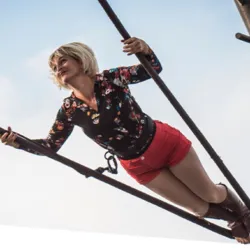 Een foto vanuit een laag perspectief van een blonde artieste in een bloemenblouse en rode korte broek, die hangt en balanceert tussen verschillende kabels die aan een monumentale metalen constructie zijn bevestigd. De foto illustreert La Promesse du Vide 