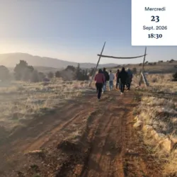 Een groep mensen loopt bij zonsopgang of zonsondergang over een zandpad richting een houten boog in een heuvelachtig natuurlandschap. Deze foto roept de reis, de beweging en de zoektocht naar evenwicht op die centraal staan in de voorstelling.