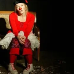Caroline Sourget, in een rood clownskostuum, zit op een stoel met een rode neus in een intieme setting tijdens een voorstelling. Ze illustreert de clownworkshop die in Laval wordt georganiseerd door de vereniging Culture et Loisirs Jean Macé.
