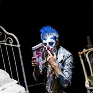 Foto van een clown die een volwassen Peter Pan uitbeeldt, geschminkt in wit en blauw, met een fotolijstje in zijn handen in een donkere kamer met een verouderde inrichting.