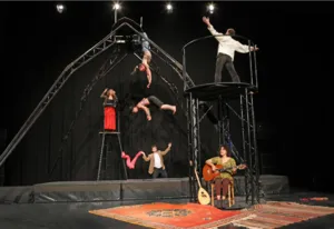 Een scène uit de show "Pour que les ailes tombent" met verschillende circusartiesten die luchtacrobatiek uitvoeren op een grote loopbrug, begeleid door een muzikant die live speelt in een warme omgeving met oosterse tapijten.