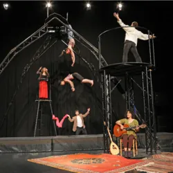 Een scène uit de show "Pour que les ailes tombent" met verschillende circusartiesten die luchtacrobatiek uitvoeren op een grote loopbrug, begeleid door een muzikant die live speelt in een warme omgeving met oosterse tapijten.