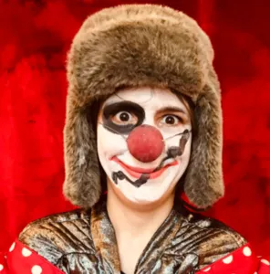 Portrait of Nicolas Cornut made up as a clown with a red nose, asymmetrical black makeup, a grey fur hat and a shiny costume, against an intense red background.
