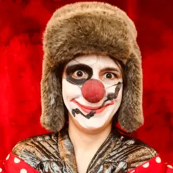 Portrait of Nicolas Cornut made up as a clown with a red nose, asymmetrical black makeup, a grey fur hat and a shiny costume, against an intense red background.