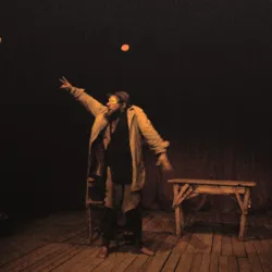 Bonaventure Gacon performs his character Boudu on a dimly lit stage. The clown, dressed in a long, worn coat, throws a ball into the air within a minimalist set consisting of a chair and a table, illustrating the dark and poetic world of