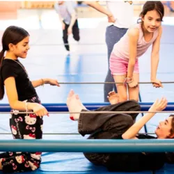 Three children are participating in a wire-dancing workshop in a gymnasium at the Fratellini Academy. Two girls stand on either side of a taut metal wire while a third, lying on the floor, works on movement and exercise techniques.