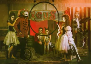 A scene of circus artists in costumes in front of a big top with an aerial hoop suspended in the foreground, illustrating the artistic universe and disciplines taught by Planet'Circus.