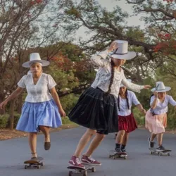 Photograph of four female skateboarders riding on a tree-lined road. They are wearing traditional clothing with wide-brimmed hats, colorful skirts, and white blouses. The image illustrates the practice of group skateboarding in a natural setting.