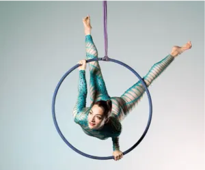 An aerial hoop artist performs within a suspended hoop against a light background. Dressed in a turquoise jumpsuit adorned with rhinestones, she executes a figure blending flexibility, core strength, and elegance, showcasing the technical skill and grace 