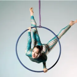 An aerial hoop artist performs within a suspended hoop against a light background. Dressed in a turquoise jumpsuit adorned with rhinestones, she executes a figure blending flexibility, core strength, and elegance, showcasing the technical skill and grace 