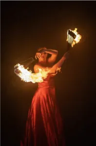An artist in a red dress performs a fire fan display in the darkness. The flames create a wide, luminous arc around the dancer, illustrating the fire arts techniques taught during the Affectus Formation professional training program.