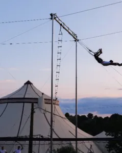 Outdoor structure of Grand Flying Trapeze installed on the CIAM site with a flyer in full flight in front of a circus tent, illustrating the professional infrastructure made available to trainees.