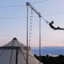 Outdoor structure of Grand Flying Trapeze installed on the CIAM site with a flyer in full flight in front of a circus tent, illustrating the professional infrastructure made available to trainees.