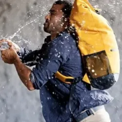An actor from the Hold up & co company wearing a yellow backpack receives a splash of water in the middle of an action, an image symbolizing the trials and tribulations experienced in the burlesque show "La Fuite" presented at the Les Toiles