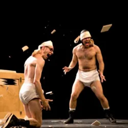 The two clowns from OKIDOK burst out laughing in the middle of a scene where pieces of wood fly through the air, illustrating the burlesque and absurd universe of Slips Inside.