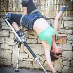 An artist equipped with a pair of fixed aluminum stilts performs an inverted acrobatic figure against a stone wall during an outdoor training session.