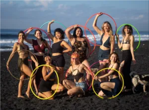 Speakers and participants of the Hoop Dream Retreat pose together on a volcanic sand beach in Bali holding multicolored hula hoops.