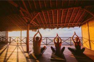 Three participants practice a mobility and meditation session on yoga mats in an open shala by the sea in Bali, at sunrise, facing the ocean.