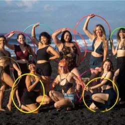 Speakers and participants of the Hoop Dream Retreat pose together on a volcanic sand beach in Bali holding multicolored hula hoops.