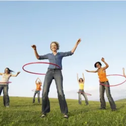 A group of adults practice hula hooping outdoors in a large green space under a clear sky, each spinning a hoop around their waist in a friendly atmosphere.
