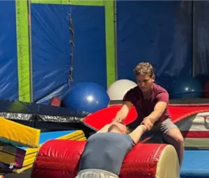 Photographie d'un exercice d'acrobatie réalisé pendant la formation TIAC dans une école de cirque. Deux stagiaires travaillent une technique d'équilibre et de renforcement sur des tapis de sécurité au sein d'un gymnase équipé de matériel circassien.