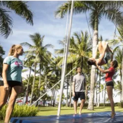 Une apprentie réalise une figure de trapèze volant sous la surveillance de deux formateurs dans une école de cirque en extérieur entourée de palmiers, illustrant la formation Animateur Cirque proposée par Club Med.