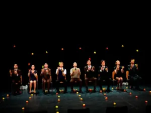 De negen artiesten van Smashed 2, die in een rij zitten, voeren tijdens de Gandini Juggling Show een gezamenlijke jongleeract uit met tientallen sinaasappels.