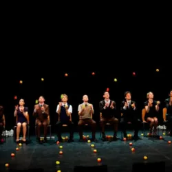 De negen artiesten van Smashed 2, die in een rij zitten, voeren tijdens de Gandini Juggling Show een gezamenlijke jongleeract uit met tientallen sinaasappels.