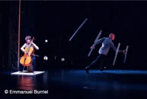 Noémi Boutin begeleidt op cello Jörg Müller bij een choreografie waarin hij jongleert met buizen die aan het podium hangen.