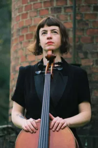 Portret van celliste Noémi Boutin met haar cello voor een bakstenen muur, presentatiefoto voor de tentoonstelling Sarabande.