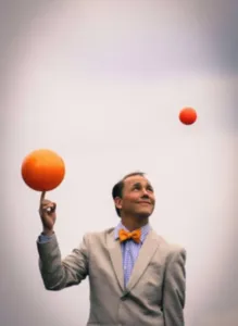 Jamie Adkins voert een jongleeract uit met verschillende oranje ballen tegen een neutrale achtergrond, ter illustratie van de Cirque symphonique-show die in april 2027 samen met het Sherbrooke Symphony Orchestra wordt gepresenteerd.