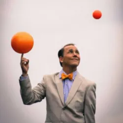 Jamie Adkins voert een jongleeract uit met verschillende oranje ballen tegen een neutrale achtergrond, ter illustratie van de Cirque symphonique-show die in april 2027 samen met het Sherbrooke Symphony Orchestra wordt gepresenteerd.