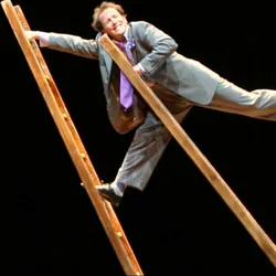 Jamie Adkins balanceert tussen twee grote houten ladders in Circus Incognitus, een circusvoorstelling die clownerie, jongleren, acrobatiek en balanceren combineert en die van 23 tot en met 26 maart 2027 te zien is in MC2 Grenoble.