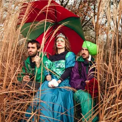 Trois artistes de la Compagnie Anoraks dans Drache Nationale, spectacle belge de jonglage, clown et cirque contemporain, installés au milieu de hautes herbes sous un grand parapluie rouge et vert, avec des balles de jonglage blanches.