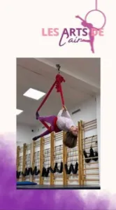 Enfant réalisant une figure suspendue à un tissu aérien rouge dans une salle de cirque, illustration des activités aériennes pour enfants proposées par Les Arts de l’Air à Nice.
