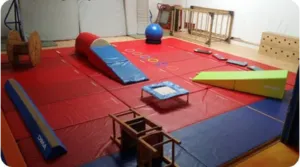 Salle de cirque Ballabulle aménagée avec tapis, mini-trampoline, modules en mousse, boule d’équilibre et matériel pour les parcours de motricité des enfants de 4 à 6 ans.