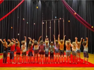 Groupe d’enfants participant à un stage d’initiation au cirque sous le chapiteau du Lido à Toulouse, les bras levés tandis que plusieurs balles de jonglage sont lancées en l’air.