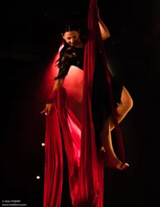 An aerial circus artist performs on a suspended red silk, illuminated by dramatic stage lighting. The performer executes an elegant aerial feat, illustrating the practice of aerial silks during a circus workshop.